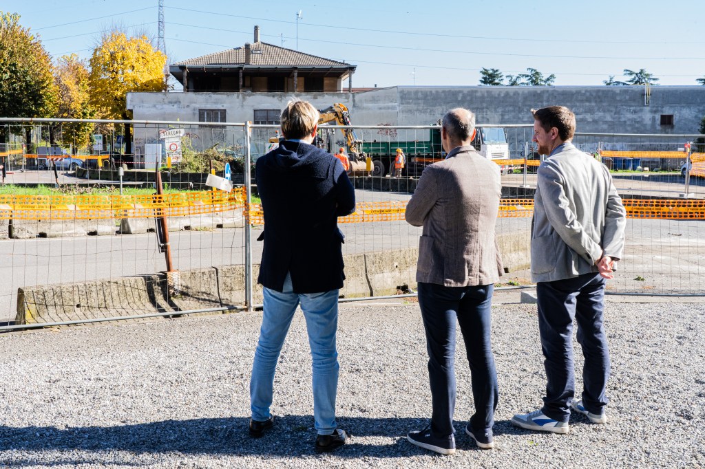Immagine che raffigura, da sinistra, il Vice Sindaco si Seregno William Viganò, che ha la delega ai Lavori Pubblici, il Sindaco di Meda, Luca Santambrogio, e il Sindaco di Seregno, Alberto Rossi, mentre osservano il cantiere da via Einaudi, verso l'imbocco di via Wagner, che è visibile sullo sfondo. 

All'interno del cantiere, da cui sono separati da grate in acciaio, si vede la vecchia rotonda fatta di barriere in New Jersey. 

Al di là di quella, in un parcheggio, sono visibili due operai con una ruspa e un altro mezzo da lavoro.