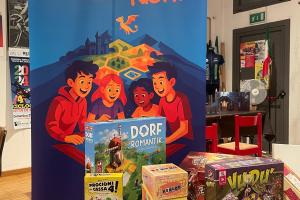 Immagine raffigurante alcuni giochi in scatola posati su un tavolo, visti frontalmente. Dietro al tavolo, un roll up raffigurante un disegno con ragazzi che giocano ad un gioco di ruolo da tavolo.