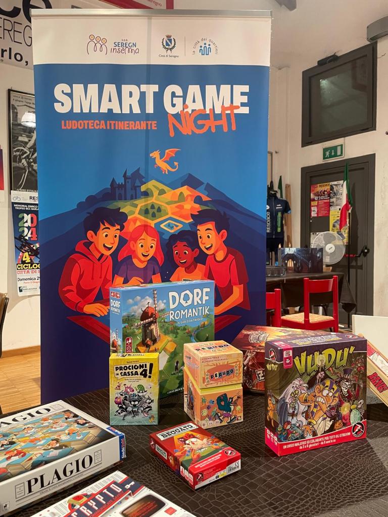 Immagine raffigurante alcuni giochi in scatola posati su un tavolo, visti frontalmente. Dietro al tavolo, un roll up raffigurante un disegno con ragazzi che giocano ad un gioco di ruolo da tavolo.