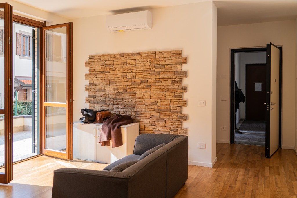 Immagine raffigurante una parte dell'area giorno, arredata con un divano e un mobiletto di uno degli appartamenti al piano terreno. Nell'immagine è presente frontalmente la porta di ingresso, aperta sull'androne del condominio, e sul lato sinistro la porta-finestra aperta sul balcone.